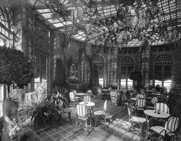 Chairs and tables are arranged throughout a dining room on the terrace of Greenbrier Hotel. The room is decorated with plants and statues and features an open roof with hanging plants below a skylight, and arched windows surrounding the round room. The Greenbrier Hotel was built in 1913.