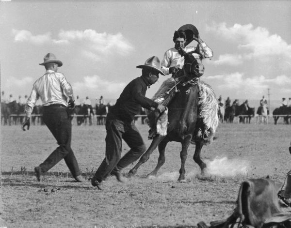 View of a "Frontier Days" Rodeo. A man holding his hat in one hand is riding a bronco while two other men run nearby. A circle of observers on horseback stand behind a barricade in the background.