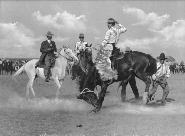 View of a "Frontier Days" Rodeo. A man is riding a bucking bronco while four other men surround him. A circle of observers on horseback are behind a barricade in the background.