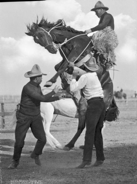 A man is riding a bucking bronco while two other men try to calm it at a "Frontier Days" rodeo.