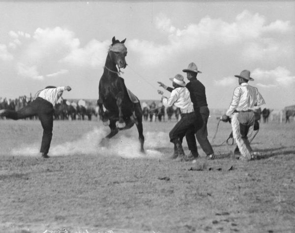 Four men attempt to saddle a bucking bronco at a "Frontier Days" rodeo.  Observers stand in the background looking on.