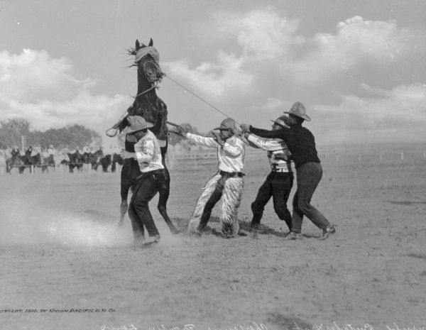 Four men approach a bucking bronco at a "Frontier Days" rodeo. A group of observers on horseback are in the background.