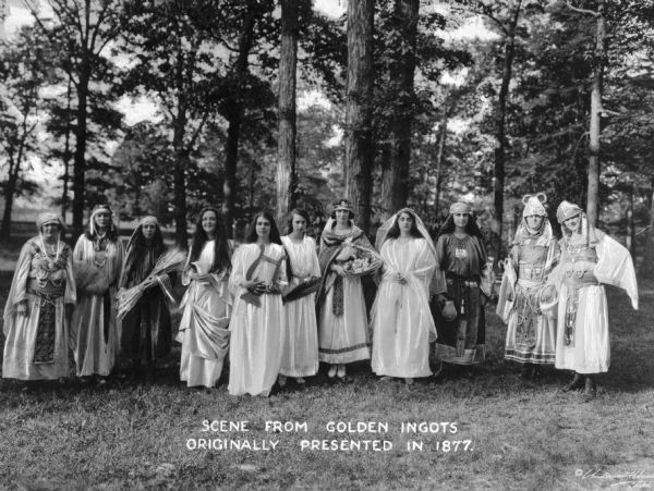 Scene from Golden Ingots at the College of Mount Saint Vincent, founded by the Sisters of Charity of New York in 1847. Costumed students act outdoors in a wooded area. Caption reads: "Scene from Golden Ingots Originally Presented in 1877."