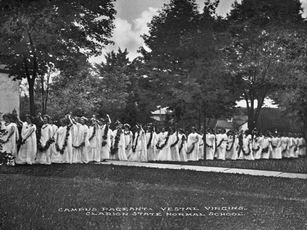 Students of Clarion State Normal School wear costumes and process past trees and school buildings during a campus pageant. Caption reads: "Campus Pageant - Vestal Virgins, Clarion State Normal School." The school was established in 1873.