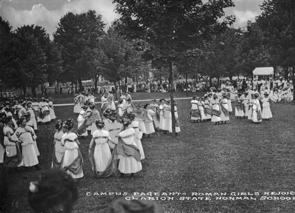 Female students in Roman costumes stand outdoors near a wooded area while putting on a pageant at Clarion State Normal School. The school was established in 1873. Caption reads: "Campus Pageant - Roman Girls Rejoice, Clarion State Normal School."