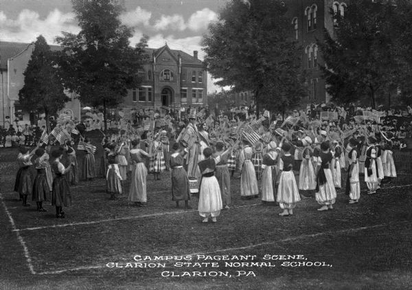 A crowd of students waving United States flags gathers on a field outside Clarion State Normal School. The school was established in 1873. Caption reads: "Campus Pageant Scene, Clarion State Normal School, Clarion, PA."