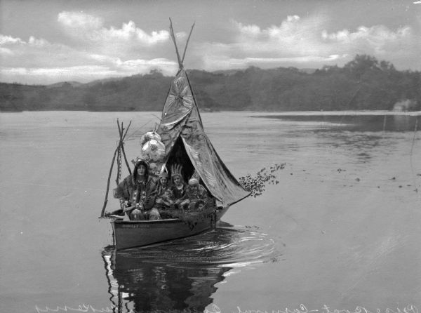 A carnival prize boat labeled "Rodney Huck" and decorated with a Native American tipi theme floats across a lake while carrying a group of costumed women and children.