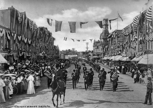 View of a parade for Old Homecoming Week. A marching band led by a man on horseback marches down a crowded commercial street, decorated with flags and bunting. A banner behind the band reads, "Old Home-Coming Week, Aug. 19-25, '06."