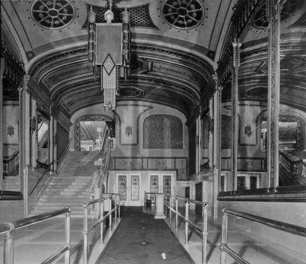 Interior of Loew's Metropolitan Theatre. The main lobby features two railings that lead toward a staircase. Mirrors line both sides of the lobby along with elegant lighting and decoration. The theater was designed by Thomas Lamb and opened on September 15, 1918.