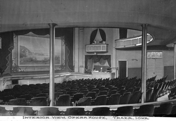 Interior of the Opera House. The view from behind the main floor seating includes the stage and the stage left balcony. In the foreground are columns supporting the mezzanine. Caption reads: "Interior View, Opera House, Traer, Iowa."