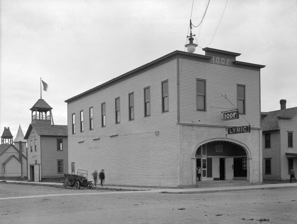 View from street of two people walking on the sidewalk near an automobile parked at the curb. They are heading toward a street corner outside the Lyric Theatre and a Grand Lodge of the Independent Order of Odd Fellows, founded in 1819. The two-story building features an arched entrance and tall windows on the upper floor. To the left of the theatre and lodge is a fire department. Signs on the facade read, "I.O.O.F." and "Lyric."