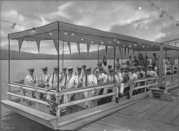 A band sits on the deck of Scott's Showboat, designed in 1932 by Ray Scott Sr., while floating near a dock on Lake Oquaga. The men are dressed in nautical costumes and hold musical instruments, and a woman in a Statue of Liberty costume sits in the center.