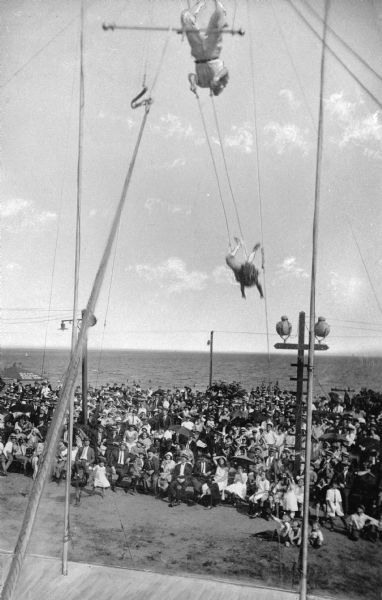 View of an acrobatic team on a trapeze. The act, performed on a boardwalk at Midland Beach by the Flying Cromwells, is observed by a large gathering of people.