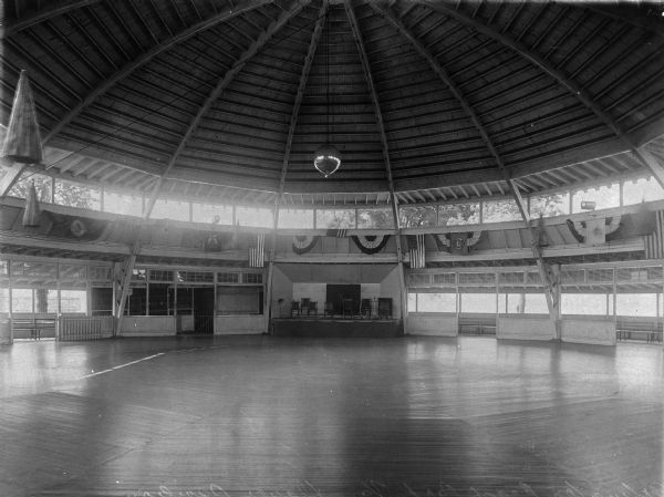 Interior of a dance pavilion. The circular building features a dance floor, stage, and disco ball. The room is decorated with bunting and American flags.