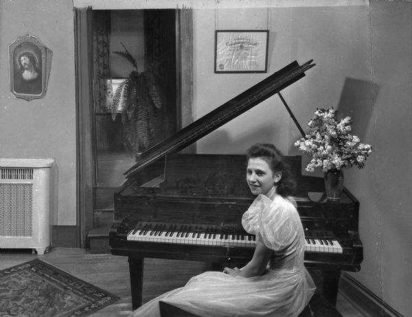 A young girl in formal dress sits before a piano. A framed certificate from the American Conservatory of Music hangs on the wall behind the piano. The American Conservatory of Music was established in 1886.