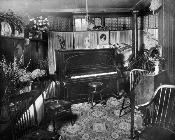 A piano stands on a carpet among chairs and decorative plants inside a music room at the Academy of Jesus and Mary. Paintings and statues stand on the piano and on a nearby shelf.
