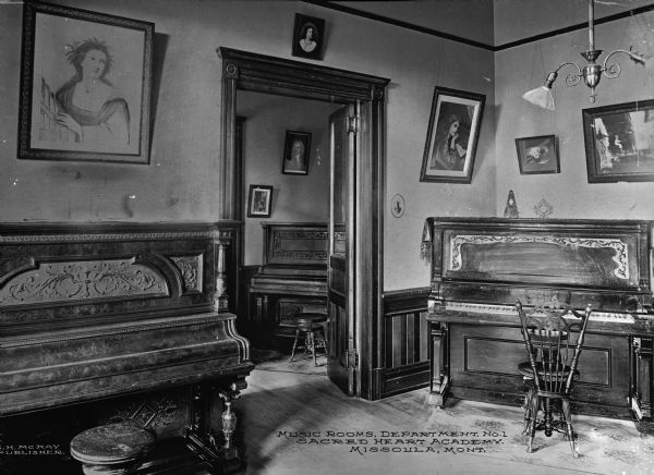 View of music rooms at Loyola Sacred Heart Academy, established in 1873.  In the first room, two pianos can be seen, and the walls are decorated with paintings. Through a doorway is another room with a piano. Caption reads: "Music Rooms, Department No. 1, Sacred Heart Academy, Missoula, Mont."