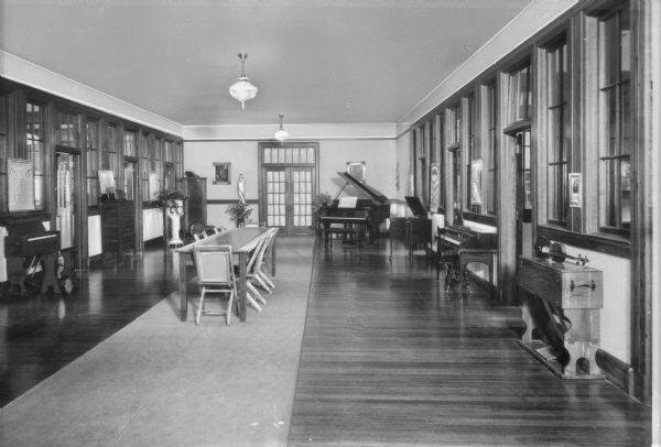 Interior of the music hall at Immaculate Heart Academy. A table and chairs stand in the center of the hall, and pianos and a violin can be seen around the room.