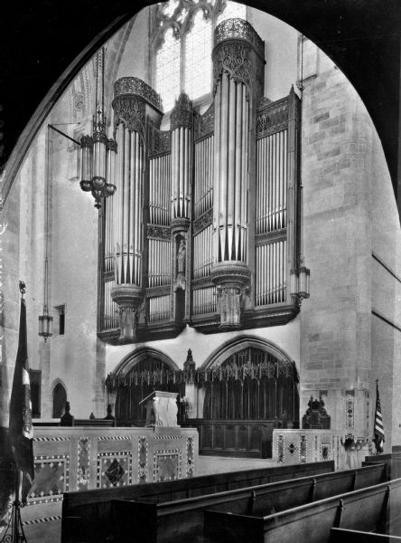 View of a large, ornate organ inside a chapel at the University of Chicago. Below the organ pipes are pews and flags. The University was founded by John D. Rockefeller in 1892.