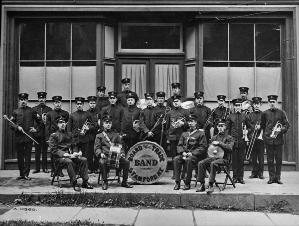 Group portrait of the Board of Trade Band. The members of the band hold instruments and stand on the sidewalk in front of a storefront.