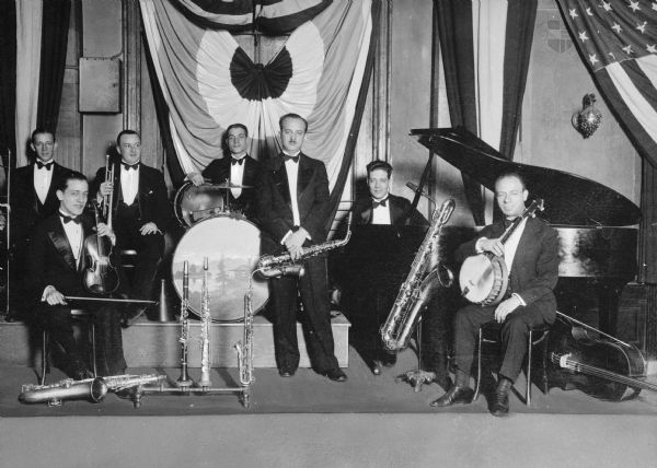 Group portrait of Simoni Martucci and Tomma's White Label Orchestra.  Members of the band hold instruments and pose before banners and decoration.
