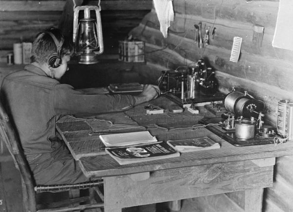 A boy scout sits at a table, wearing headphones, to send and receive radio messages. A kerosene lamp hangs behind him.