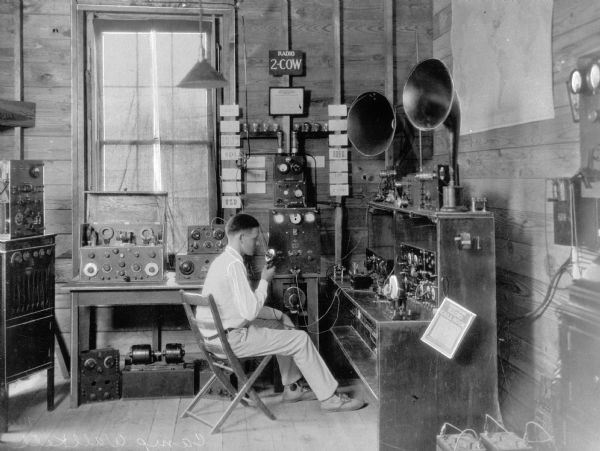 A boy sits at a wooden desk to send radio calls at Camp Wallkill.  Radio equipment is arranged on the desk and surrounding tables, a radio call book can be seen on a stand on the right, and a map hangs on the wall.