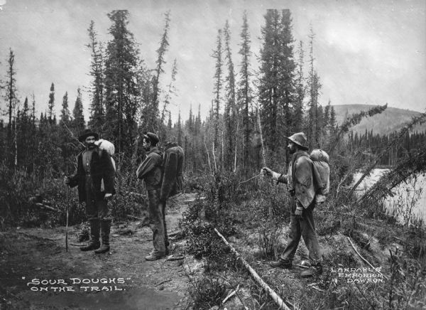 View of miners walking through a wooded area. Caption reads, "'Sour Doughs' On The Trail."