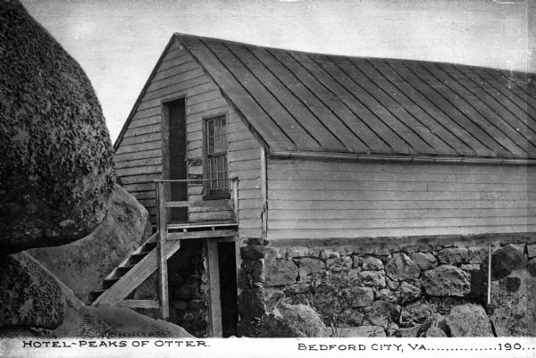 Exterior of Peaks of Otter Lodge. The wooden shelter, built into the surrounding rock, features a doorway in the stone, ground floor, and one in the wooden upper story. Caption reads: "Hotel — Peaks of Otter. Bedford City, VA....190..."