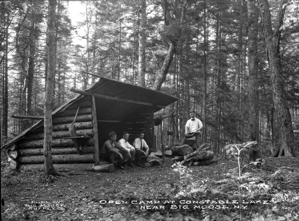 View of four men at Constable Lake Campsite. Three men sit inside a lean-to shelter, and one man stands nearby, holding a pan. Caption reads: "Open Camp at Constable Lake Near Big Moose, N.Y."