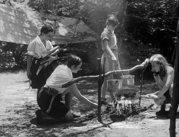 View of four girls fixing dinner over a campfire. Two pots hang from a spit over the fire and a girl carries an armful of logs. Behind them, a tent can be seen in a wooded area.
