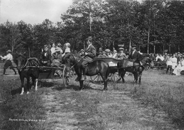 Boys ride ponies and travel in horse-drawn carriages through a field at Buck Hill Falls, a private resort community in the Pocono Mountains.   Behind them, a large crowd of people sit at picnic tables near signs that advertise a restaurant and ice cream. Buck Hill Falls was founded in 1901.