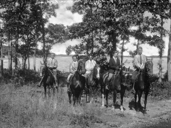 View of men and women horseback riding in the woods at Peconic Lodge.  A large body of water is in the background.