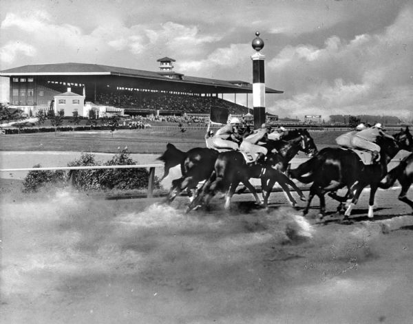 Jockeys on horseback pass a striped marker on the dirt race track at Arlington Park. In the background the grandstand holds numerous spectators. Arlington Park opened on October 13, 1927.