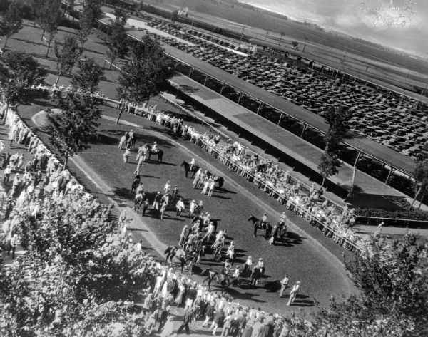 Jockeys exercise their horses in the paddock at Arlington Park while spectators look on. The elevated view shows a crowded parking lot at right. Arlington Park opened on October 13, 1927.