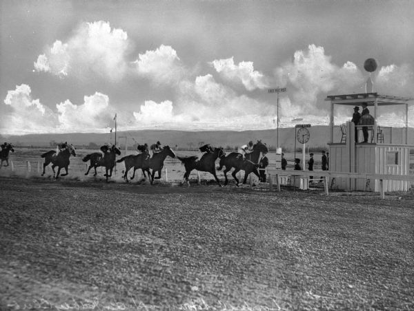 View of a horse race at the Lower California Jockey Club in Tijuana, Mexico. Two men stand in a shelter, keeping time, while others watch the horses race past the "first past post" from the sidelines. A sign shows that the next race begins at 2:30.