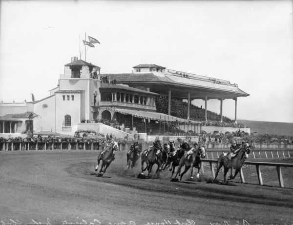 Horses race past on the track at Aqua Caliente Jockey Club while spectators look on from the grandstand. The club opened in 1929 and was located in Tijuana, Mexico.