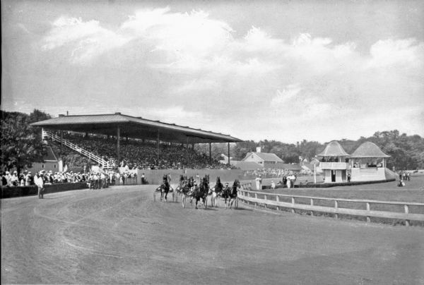 Horses harnessed to sulkies pass the judge's stand at Goshen Historic Track as spectators look on from the grandstand and sidelines. Goshen Historic Track opened in 1838 and is known as the world's oldest harness track. The grandstand was constructed in 1911.