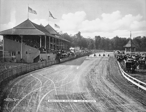 Elevated view of racing trotters lined up on the track at Lima Driving Park as spectators look on from the grandstand. Automobiles are parked near the judge's stand to the right. Lima Driving Park was established in 1903. Caption reads: "Driving Park, Lima, Ohio."