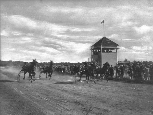 Harnessed horses race past the judge's stand while spectators watch from the sidelines at The Finish, a dirt racetrack.