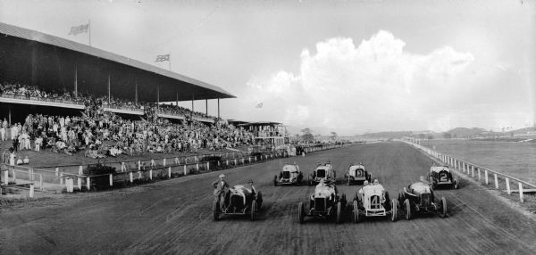Drivers and passengers stand on the dirt track at Oriental Park Racetrack before an automobile race. On the left is a grandstand with spectator. Oriental Park Racetrack was founded in 1915 in Havana, Cuba.