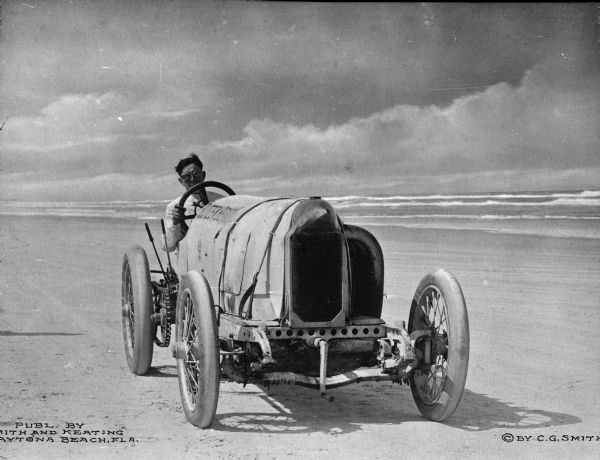 A man wearing goggles drives a Blitzen Benz automobile on the sand along a shoreline. The Blitzen Benz was first built in 1909 by Benz & Cie.