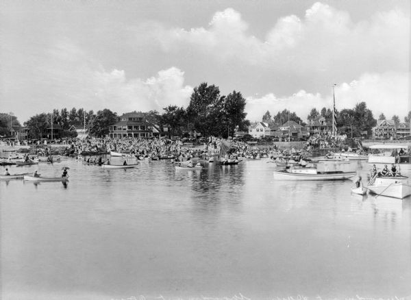 Several varieties of boats crowd Woodmont River and crowds gather along the shoreline. Houses are in the background and automobiles are parked along the roadside.