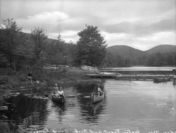 Girls canoe near a dock at Camp Lenoloc as additional girls watch from the shoreline. Trees and other foliage line the water and mountains are in the background.