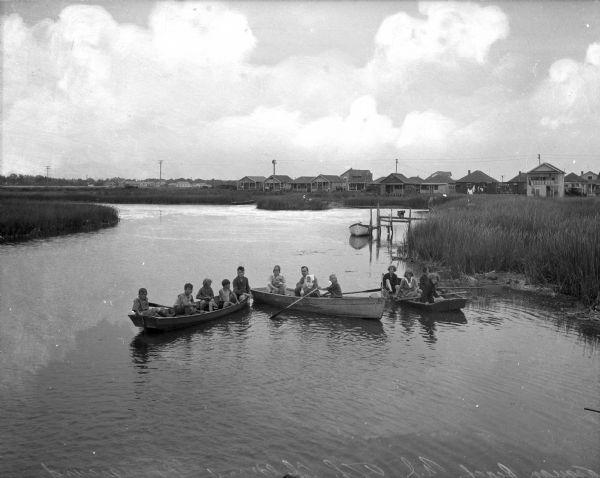 Elevated view of a group of children, infants, and adults sitting in rowboats in the water at Manasquan Beach. In the background are rows of dwellings, and another boat is tied to a dock near the marshy shoreline.
