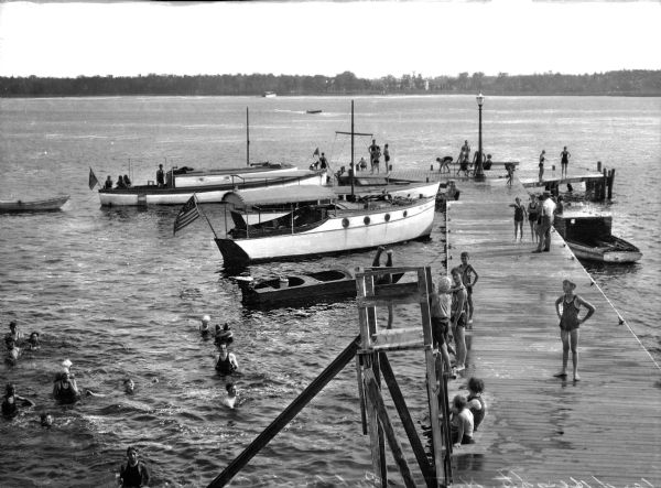 Elevated view of adults and children wearing bathing costumes standing on a wooden dock crowded with several varieties of boats. People are swimming in the water on the left. In the background is a wooded shoreline.