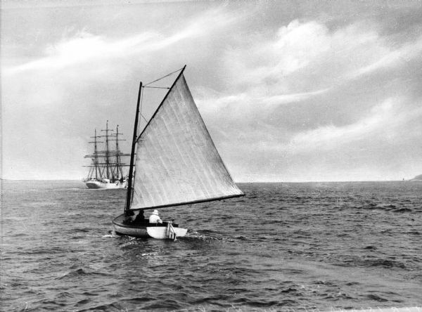View of a family sailing in Dering Harbor. The sailboat is carrying a man, woman, and girl. In the background is a small ship.