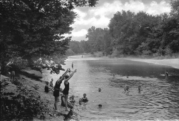 A group of people bathe at Miller's Swimming Hole at Elk Springs.  Three people prepare to dive into the water while others swim or sit on the rocks.