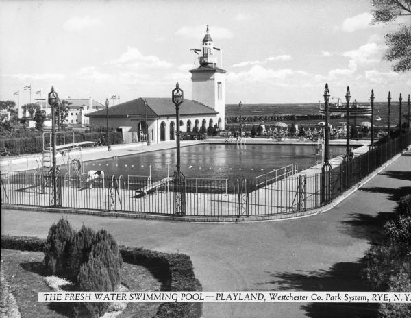View of an outdoor freshwater swimming pool, which opened in 1928. Two people lean over the fence to  look into the pool and, behind them, a building with a tower leads to the boardwalk and cafeteria. In the background, a ship is docked at a pier in Playland Lake. Caption reads: "Freshwater Swimming Pool -- Playland, Westchester Co. Park System, Rye, N.Y."