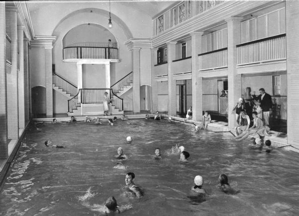 Children and adults swimming in a swimming pool inside Bedford Springs Hotel. Other people are sitting and standing near the edge. An elaborate staircase on the far wall leads to the balconies surrounding the pool.